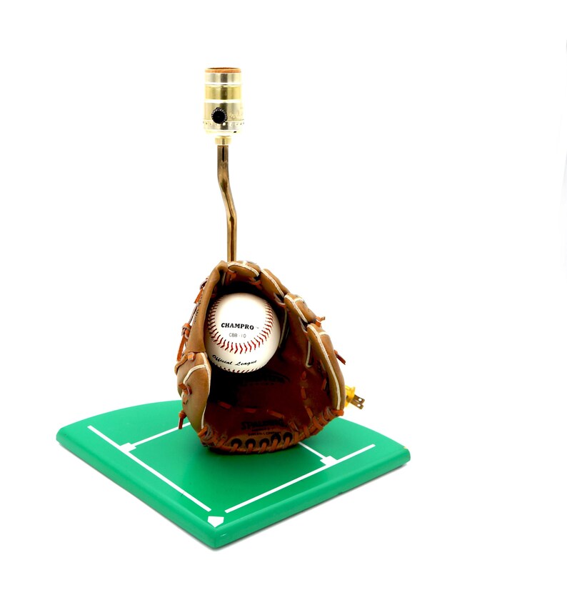 K&ouml;nnte beinhalten: Eine Baseball-Lampe mit einem Baseballhandschuh, der einen Baseball h&auml;lt. Der Baseball tr&auml;gt den Text "CHAMPRO Official League". Die Lampe steht auf einer gr&uuml;nen, rautenf&ouml;rmigen Basis.