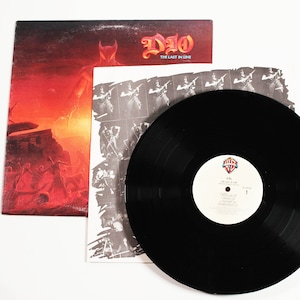 May include: A black vinyl record with a white label that says "DIO - The Last in Line" and a picture of a man in a horned helmet. The record is on top of a white inner sleeve with a black and white photo of a man playing a guitar. The record is on top of a red and orange album cover with a picture of a man in a horned helmet.