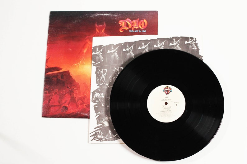 May include: A black vinyl record with a white label that says "DIO - The Last in Line" and a picture of a man in a horned helmet. The record is on top of a white inner sleeve with a black and white photo of a man playing a guitar. The record is on top of a red and orange album cover with a picture of a man in a horned helmet.