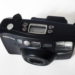 A dark gray Pentax IQZoom 140 35mm film camera is angled from the top right, showing the front, top, and right side with its lens, flash, and LCD screen.