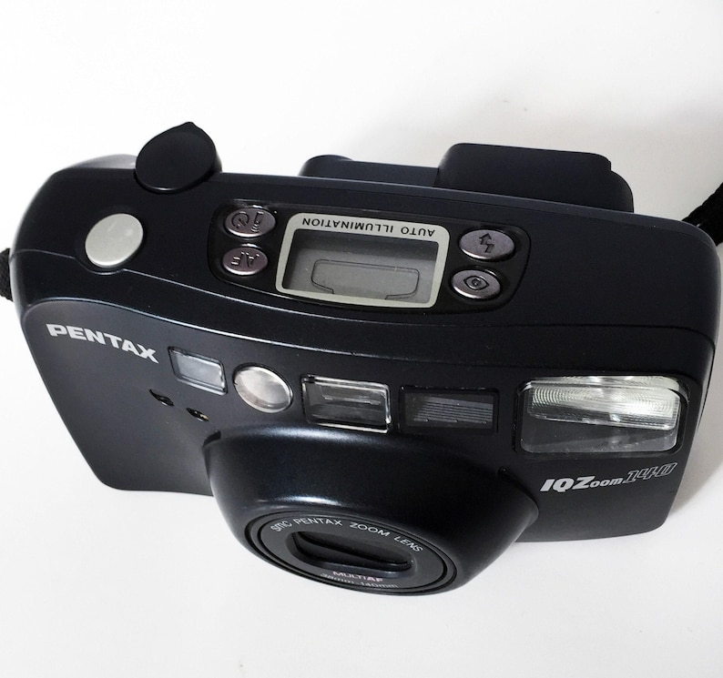 A dark gray Pentax IQZoom 140 35mm film camera is angled from the top right, showing the front, top, and right side with its lens, flash, and LCD screen.