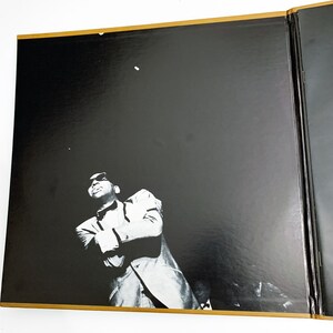 Pu&ograve; includere: Fotografia in bianco e nero di un uomo in giacca e cravatta, che guarda in alto. L'immagine &egrave; su uno sfondo nero con un bordo dorato. L'uomo &egrave; nella parte inferiore dell'immagine, con la maggior parte dell'immagine che &egrave; lo sfondo nero.