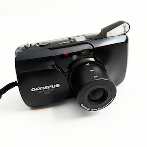 May include: A black Olympus Stylus Zoom camera with a built-in flash. The lens has a zoom range of 35-70mm.