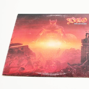 May include: A vinyl record album cover featuring a red and orange illustration of a large, demonic figure with horns and a glowing red eye. The figure stands in front of a fiery, apocalyptic landscape. The album title "DIO - THE LAST IN LINE" is printed in black at the top right corner.