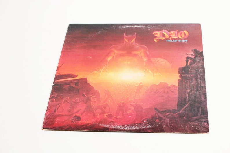 May include: A vinyl record album cover featuring a red and orange illustration of a large, demonic figure with horns and a glowing red eye. The figure stands in front of a fiery, apocalyptic landscape. The album title "DIO - THE LAST IN LINE" is printed in black at the top right corner.