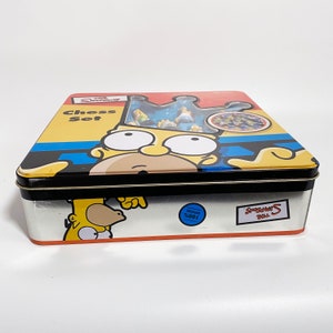 May include: A yellow metal tin box with a Simpsons Chess Set design. The box features a cartoon image of Homer Simpson wearing a crown. The text "The Simpsons Chess Set" is printed on the box.