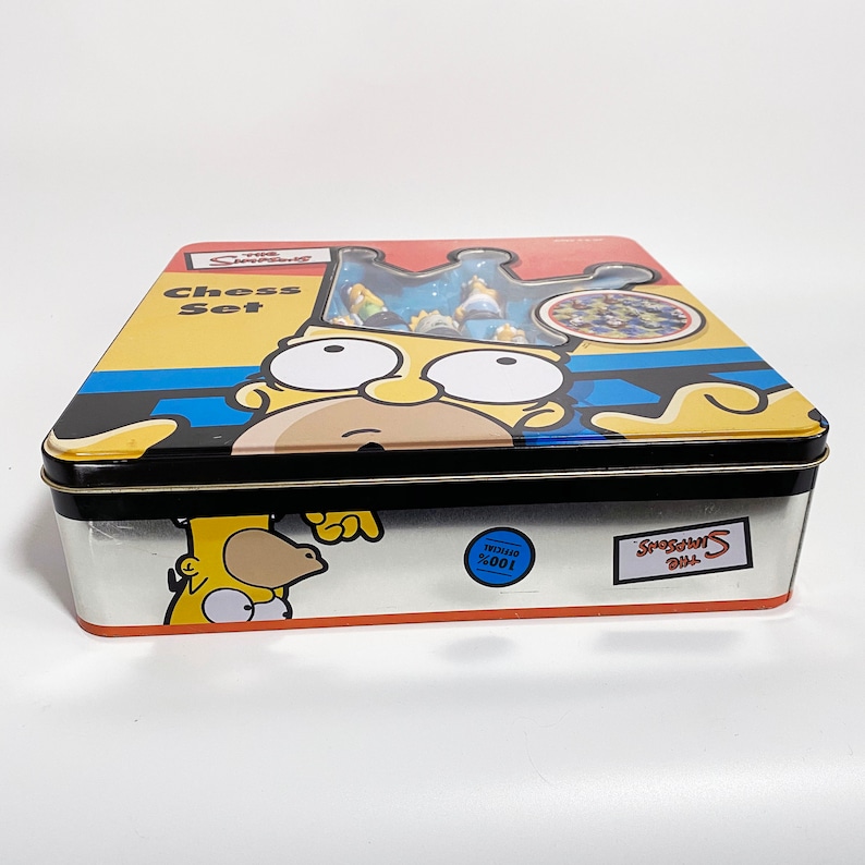 May include: A yellow metal tin box with a Simpsons Chess Set design. The box features a cartoon image of Homer Simpson wearing a crown. The text "The Simpsons Chess Set" is printed on the box.
