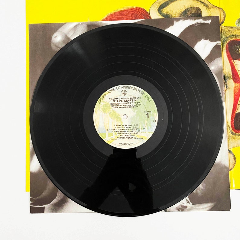 May include: Black vinyl record with a white label featuring the text "Burbank Home of Warner Bros Records", "William E. Meulen Presents", "Steve Martin", "Comedy Is Not Pretty", "Produced by William E. Meulen", "Aspen Recording Society", and "1 0".