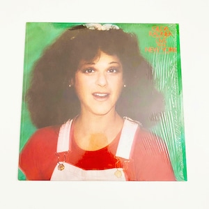 May include: A vinyl record album cover featuring a woman with curly brown hair wearing a red shirt and white overalls. The album title is "CILLA BLACKNER LIVE AT THE NEW YORK", with a green background.
