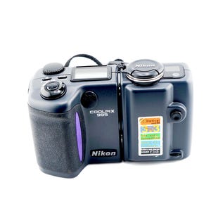 May include: A dark gray Nikon Coolpix 995 digital camera with a swivel lens design. The camera features a textured grip, a top-mounted shutter button, a lens cap labeled Nikon, and a multi-colored sticker on the side detailing technical specifications.