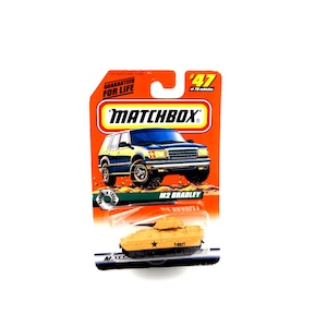 May include: A Matchbox toy set featuring an M2 Bradley tank and a blue SUV. The packaging is orange with the Matchbox logo and the text "GUARANTEED FOR LIFE". The tank is tan with a black star and the number "1-0927".