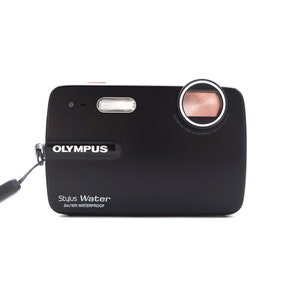 May include: A black Olympus Stylus Water camera with a silver lens and a silver button on the top. The camera is waterproof to 3 meters or 10 feet. The text "Olympus Stylus Water 3m/10ft Waterproof" is printed on the front of the camera.