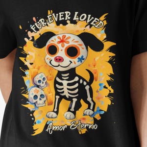 May include: Black V-neck t-shirt featuring a Day of the Dead-themed dog illustration. The dog has a skeleton body and a decorated skull face. Text includes "Fur Ever Loved" and "Amor Eterno" on a yellow and orange background.