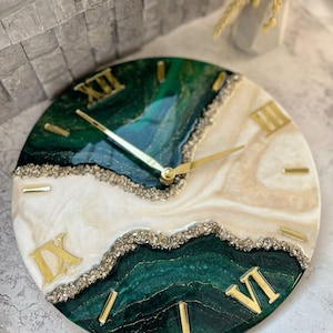 Può includere: Orologio da parete rotondo dal design unico. Il quadrante presenta una miscela di resina verde, bianca e beige, con numeri romani e lancette dorate. Il bordo dell'orologio è adornato da un bordo strutturato e scintillante.
