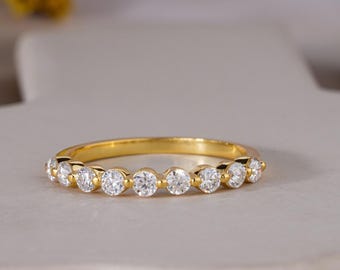 May include: A gold band ring with a series of round, clear gemstones set across the top. The ring is displayed on a light gray surface, with yellow flowers in the background. The ring is a classic design.
