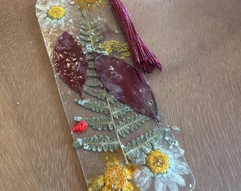 May include: Clear resin bookmark with embedded dried flowers and leaves. The bookmark features a burgundy tassel and a small loop for easy placement. The design includes yellow and white daisies, fern leaves, and burgundy leaves.