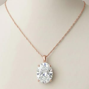 Puede incluir: Un collar de oro rosa con un gran colgante de diamante de talla ovalada. El diamante está engastado en un bisel de oro rosa y cuelga de una delicada cadena de oro rosa. El collar se exhibe en un maniquí blanco.