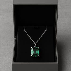 Könnte beinhalten: Eine silberne Halskette mit einem großen, smaragdgeschliffenen, grünen Edelsteinanhänger, präsentiert in einer offenen grauen Schmuckschatulle mit schwarzem Innenraum. Die Halskette ist vor dem schwarzen Hintergrund zentriert.