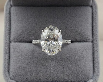 May include: A silver-coloured ring featuring a large, oval-cut clear gemstone held by six prongs. The band is thin and encrusted with small, round, clear stones along the visible portion of the shank, creating a sparkling, reflective appearance.