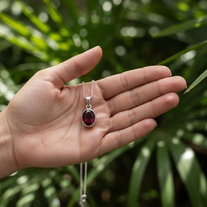 May include: A silver pendant necklace featuring an oval, deep red gemstone. The pendant is suspended from a delicate silver chain. The background is a blurred view of green foliage.
