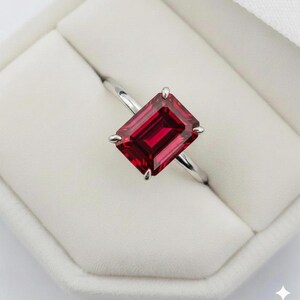 May include: A silver ring featuring a large, rectangular, deep red gemstone. The gemstone is set with four prongs, and the ring is presented in a white jewelry box. The ring's band is thin and polished.