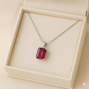 May include: A silver necklace with a rectangular, deep red gemstone pendant. The pendant is set in a silver frame and hangs from a delicate silver chain. The necklace is presented in an off-white jewelry box.
