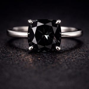 Könnte beinhalten: Ein silberner Ring mit einem großen, quadratisch geschliffenen schwarzen Edelstein. Der Ring hat ein schlichtes Band und vier Krappen, die den dunklen Stein halten. Der Ring ist vor einem dunklen Hintergrund platziert.