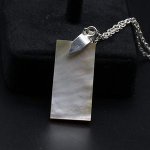 Puede incluir: Un collar con colgante rectangular con una cara de nácar. El colgante está unido a una cadena de plata. El colgante mide aproximadamente 2,5 cm de largo y 1,3 cm de ancho. El collar se muestra sobre una superficie negra.