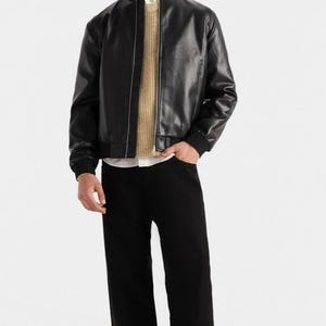 Puede incluir: Chaqueta bomber de cuero negro con cuello y puños de canalé. La chaqueta se lleva sobre un suéter beige y una camisa blanca con cuello. La persona lleva pantalones negros y zapatos negros. El conjunto es de estilo moderno e informal.