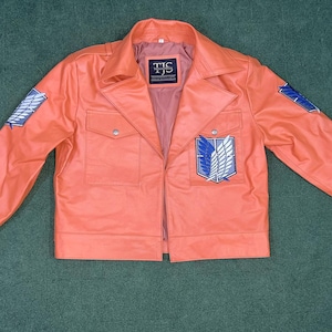 May include: Coral-colored jacket with a collared neckline, two chest pockets, and long sleeves. Features a patch on each sleeve and a larger emblem on the chest. The jacket has the text "TJS" on the inner label.