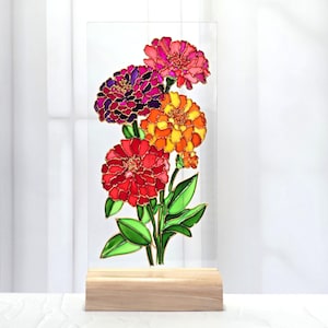 以下が含まれることがあります： 赤、オレンジ、ピンク、紫の色合いの4輪のジニアの花と緑の葉が描かれた、ステンドグラス風の絵が施された長方形の透明なガラスパネル。明るい色の木製の長方形の台座に取り付けられています。