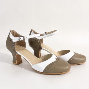 May include: A pair of two-tone taupe and white heels featuring a rounded toe, a cutout side design, and a thin ankle strap with a silver-toned metal buckle. The shoes include decorative brogue-style perforations along the toe and heel sections.
