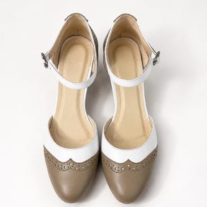 May include: A pair of two-tone Mary Jane shoes featuring a white upper section and a taupe-colored rounded toe cap. The shoes include a decorative brogue-style perforated trim along the seam, a thin ankle strap with a metal buckle, and a beige insole.