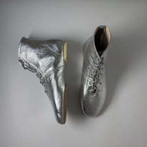 Pode incluir: Um par de botas de cano alto com acabamento em prata metálica. As botas apresentam um fecho frontal com atacadores prateados a condizer, uma biqueira arredondada e uma sola plana de cor castanha clara. O material tem uma textura.