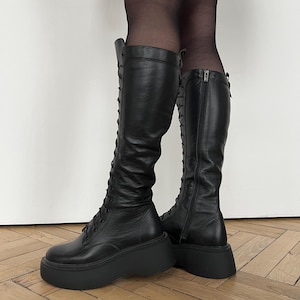 May include: Black knee-high boots featuring a lace-up front design with thin black laces, a rounded toe, and a thick, chunky platform sole. The boots include a vertical zipper closure on the inner side and a smooth, matte leather-like finish.