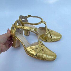 May include: A pair of gold-colored, heeled sandals. The sandals have a woven design across the toes and a T-strap that fastens around the ankle. The heels are approximately 2 inches (5 cm) high.