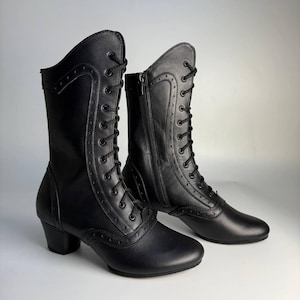 May include: Black leather boots with a vintage aesthetic. These boots have a lace-up front, a side zip, and a small heel. The design includes decorative stitching and a classic shape, suitable for different styles. The boots are approximately 20 cm high.