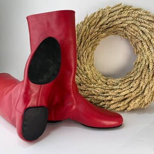 May include: A pair of bright red leather mid-calf boots featuring a smooth finish, a rounded toe, and a distinct black split sole design on the bottom. The boots have a simple pull-on construction with no visible zippers or laces, showcasing a clean silhouette.