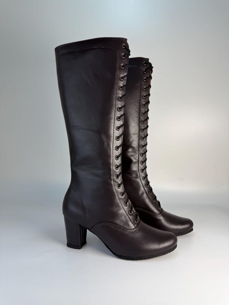 May include: A pair of dark brown, knee-high boots with a lace-up design. The boots have a rounded toe, a block heel, and a smooth leather-like texture. The laces are black, and the boots are set against a light gray background.