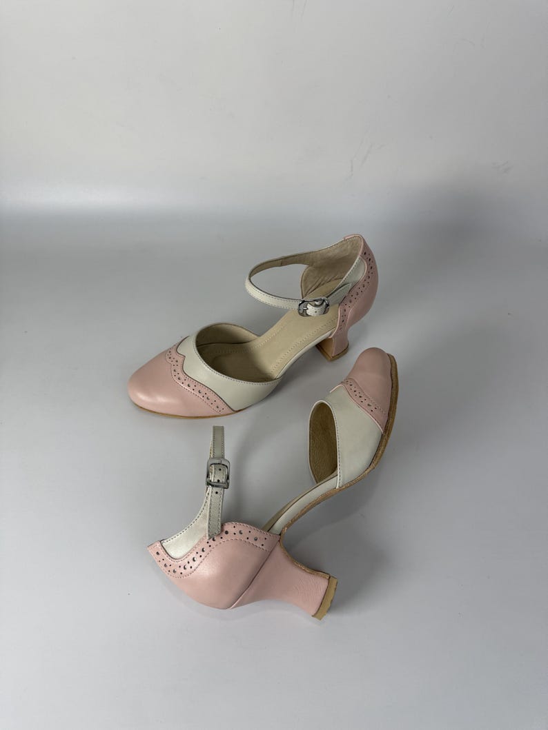 Op de afbeelding: Een paar tweekleurige Mary Jane-schoenen in lichtroze en cr&egrave;me. Deze schoenen hebben een ronde neus, een lage blokhak en een enkelbandje met gesp. Het ontwerp bevat decoratieve perforaties in brogue-stijl langs de naden van de schoen.