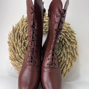 May include: A pair of mid-calf brown boots featuring a lace-up front with dark brown laces, decorative brogue-style perforations along the seams, a rounded toe, and a side zipper closure. The boots have a smooth finish and a solid black sole.