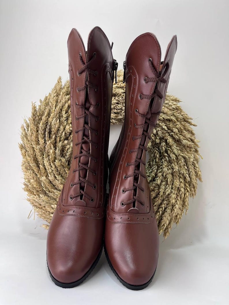 May include: A pair of mid-calf brown boots featuring a lace-up front with dark brown laces, decorative brogue-style perforations along the seams, a rounded toe, and a side zipper closure. The boots have a smooth finish and a solid black sole.