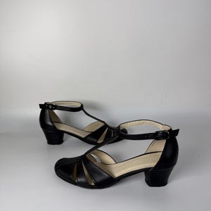 May include: A pair of black leather T-strap shoes featuring a rounded toe, cut-out side panels, an adjustable ankle strap with a small metal buckle, and a low block heel. The interior lining is a light beige colour, providing a contrast to the exterior.