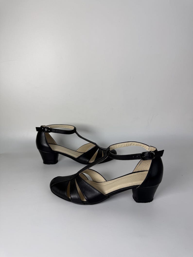 May include: A pair of black leather T-strap shoes featuring a rounded toe, cut-out side panels, an adjustable ankle strap with a small metal buckle, and a low block heel. The interior lining is a light beige colour, providing a contrast to the exterior.