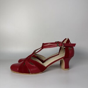 Puede incluir: Un par de zapatos rojos con correa en T que presentan una combinaci&oacute;n de texturas lisas y tipo ante. El calzado incluye una puntera redondeada, un tac&oacute;n de bloque de altura media y una correa de tobillo ajustable con una peque&ntilde;a hebilla de metal dorado.