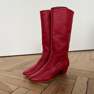 Pode incluir: Um par de botas altas de couro vermelho. As botas t&ecirc;m uma textura lisa, uma ponta arredondada e um salto pequeno. As botas s&atilde;o at&eacute; o joelho e est&atilde;o sobre um ch&atilde;o de madeira. O fundo &eacute; uma parede branca lisa.