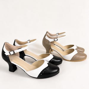 May include: Two pairs of Mary Jane heels with a rounded toe and a low block heel. One pair is black and white, and the other is tan and white. Both feature a buckled ankle strap, a cutout side design, and decorative brogue-style perforations.