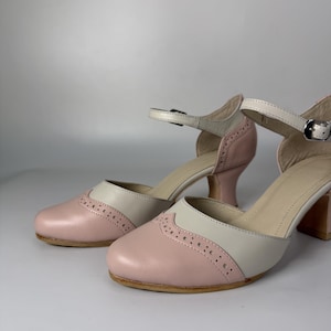 Op de afbeelding: Een paar tweekleurige schoenen met een zachtroze ronde neus voorzien van decoratieve brogue-perforaties en een lichtbeige bovenzijde. De schoenen hebben een verstelbaar enkelbandje met een zilverkleurige metalen gesp en een blokhak.