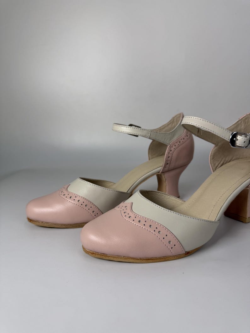 Op de afbeelding: Een paar tweekleurige schoenen met een zachtroze ronde neus voorzien van decoratieve brogue-perforaties en een lichtbeige bovenzijde. De schoenen hebben een verstelbaar enkelbandje met een zilverkleurige metalen gesp en een blokhak.