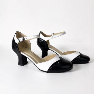 May include: A pair of black and white two-tone shoes with a rounded toe, a mid-height block heel, and an ankle strap featuring a silver-toned metal buckle. The design includes decorative perforations along the wingtip-style toe cap seam.